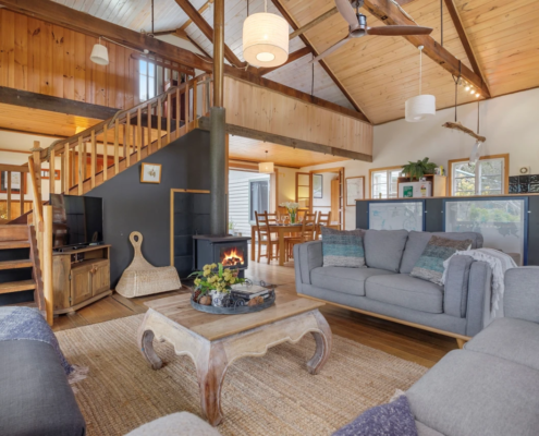 Main Living Area: An open plan living room with wood heater, timber beams, ceiling fan and comfy sofas centered around a coffee table.